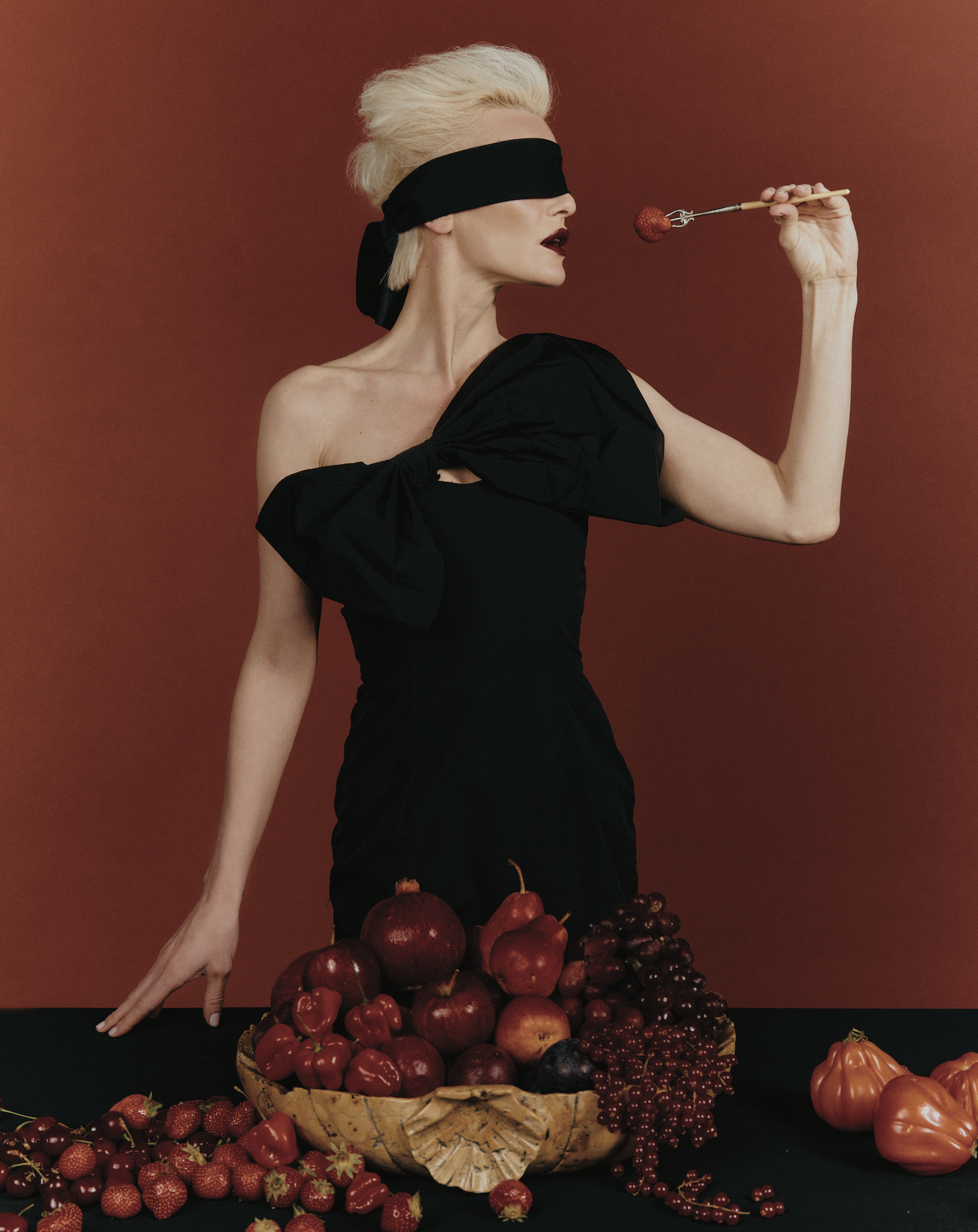 Woman in a black dress, blindfolded, holding a spoon against a deep red backdrop with fruit arranged in front of her